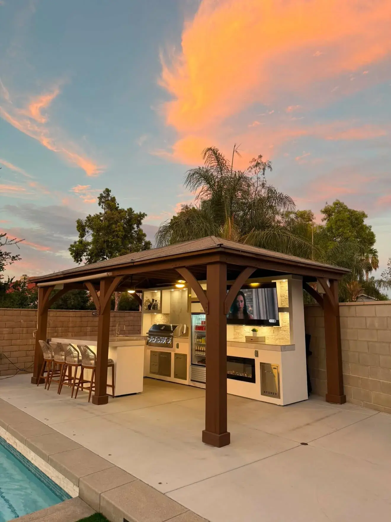 Timber pergola sheltering an outdoor kitchen beside a pool at sunset