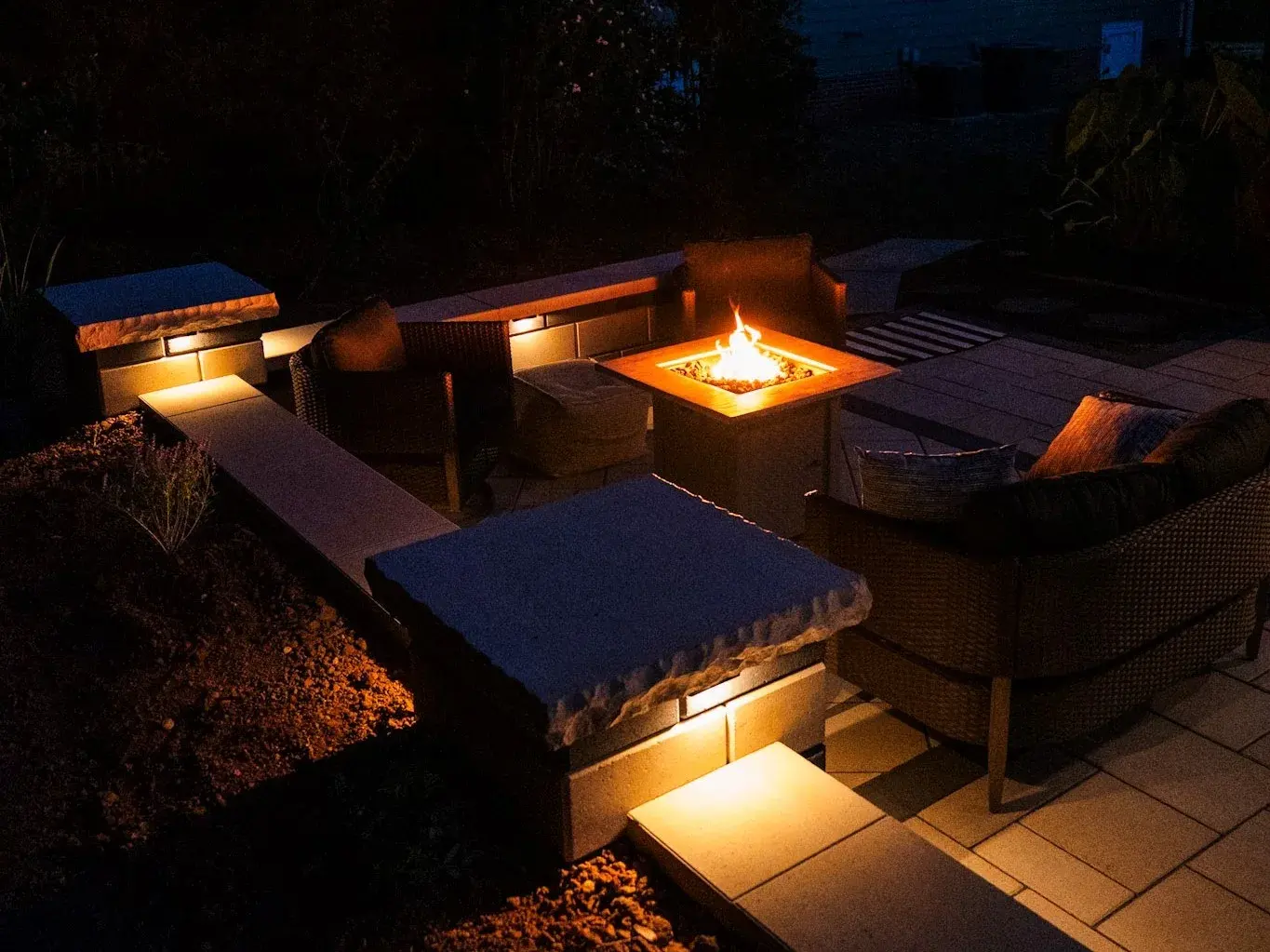 Paver patio at dusk with built-in seat walls, cap lighting, and a glowing fire table
